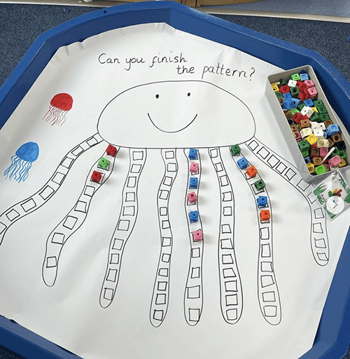 A simple octopus drawn onto a large piece of paper placed inside a tuff tray. The tentacles have segments for placing coloured blocks, with the words 'can you finish the pattern' written above the octopus
