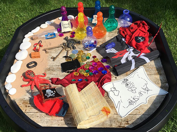 A tuff tray filled with pirate-related bits and pieces like jolly roger, a couple of maps, plastic gems and coins and 'pirate potions'