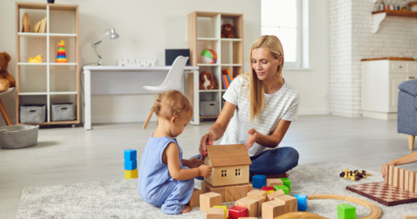 A woman building a toy wooden house with a blonde child, surrounded by toy building blocks like the best early years practitioners
