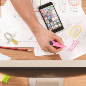 A man's arms reaching down and writing on a pile of papers, with a computer screen at the foot of the image with post-it notes and a pen under it to show marketing planning