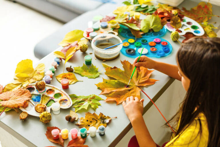 A young child painting a leaf at a small table surrounded by crafts and other pieces from nature