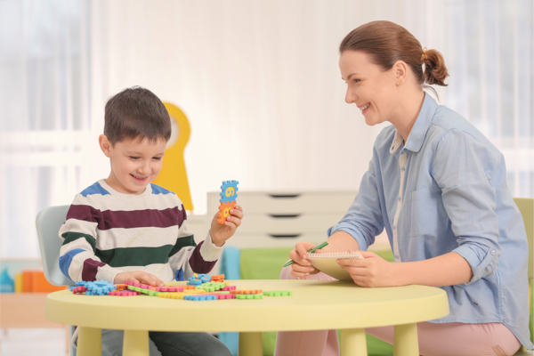 A woman and a child at a table, the child is playing with interlocking squares and the woman is prepped with a notepad