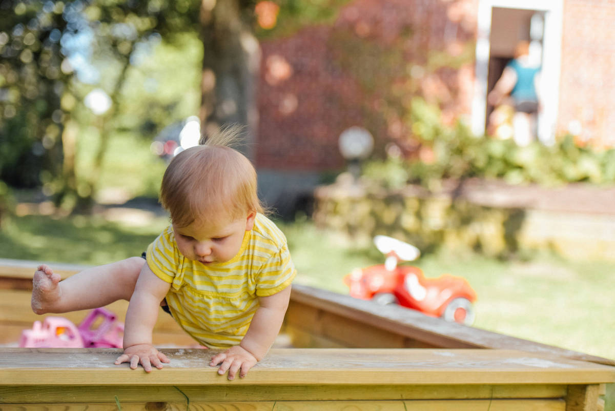 A child climbing out of a wooden sandpit on a sunny day. A red trike is visible in the background