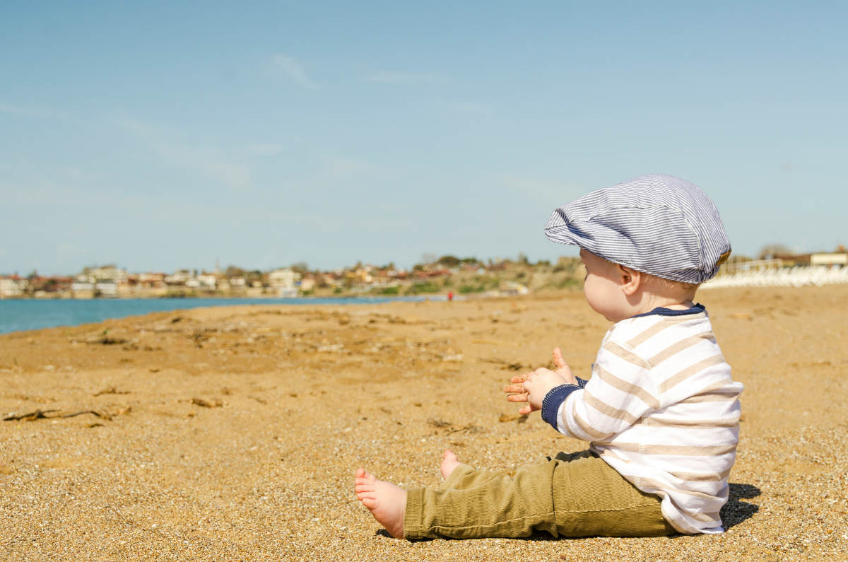 A child with a flat cap on sat on a beach playing with sand