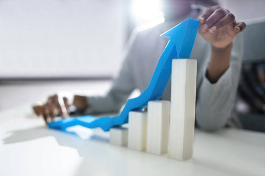 A 3D representation of a bar and line graph, rendered in the real world with someone's hand pointing to the tip of the line graph, trending upward