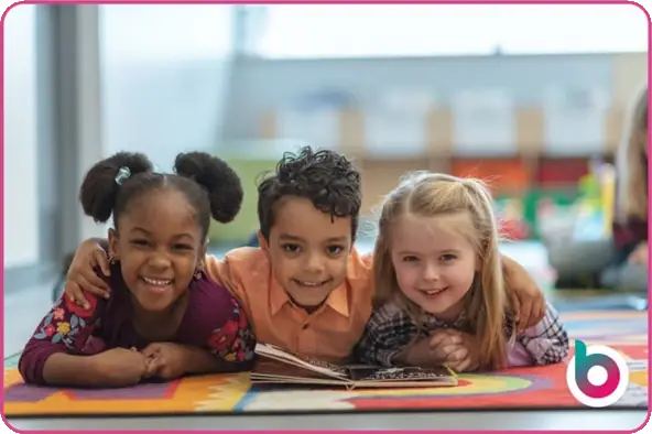 three children lying on the floor looking forwards and smiling behind an open book